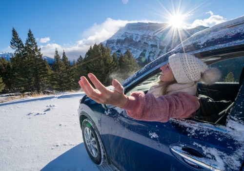 Mulher jovem no carro numa estrada de montanha esticando os braços para fora da janela. Paisagem de montanha no inverno com neve a cobrir a estrada e pinheiros. 